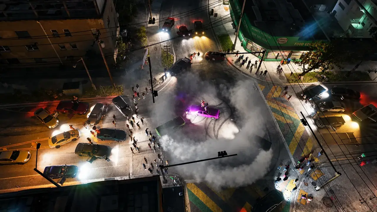 A crowd surrounds cars and tire smoke at a nighttime street takeover in GTA VI, viewed from above.